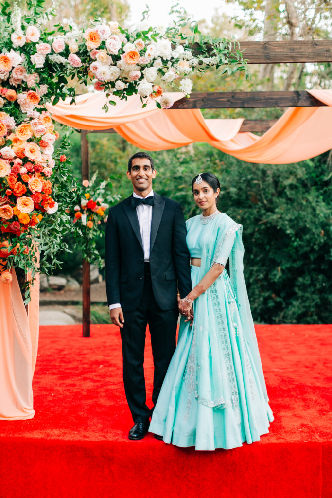 bride in teal wedding saree and groom in black suit portrait shot in front of mandap