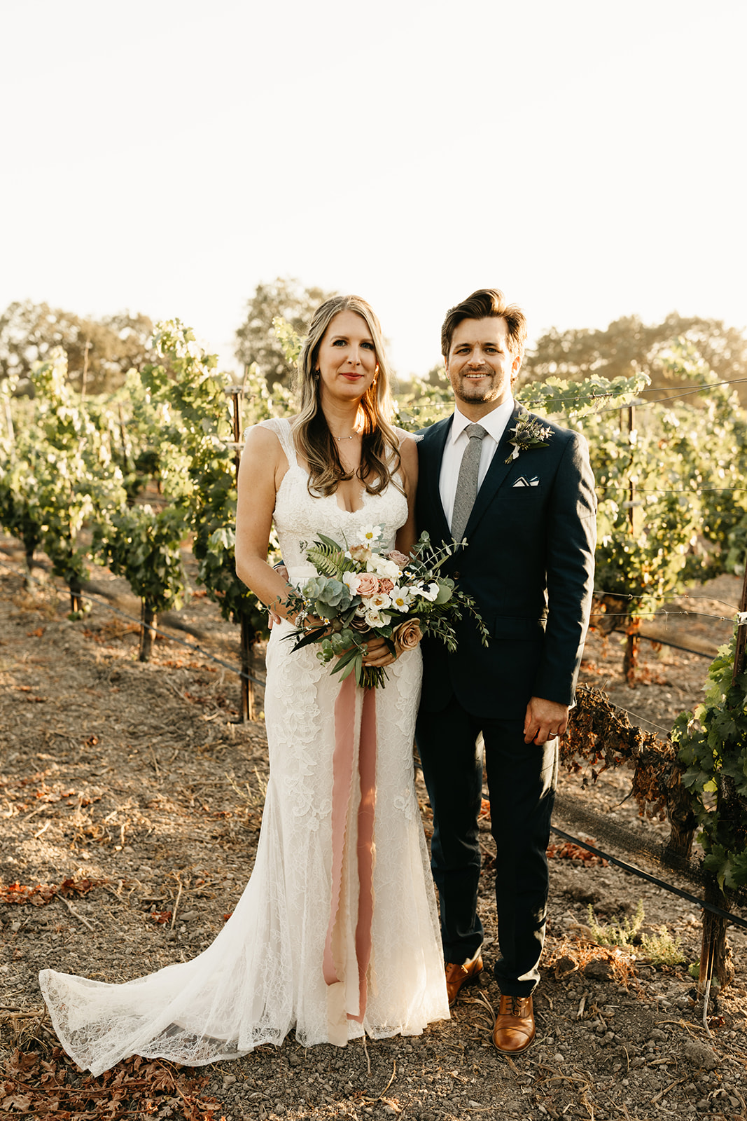 bride in lace dress and groom in dark blue suit take sunset portrait shots in vineyard at Roblar Winery