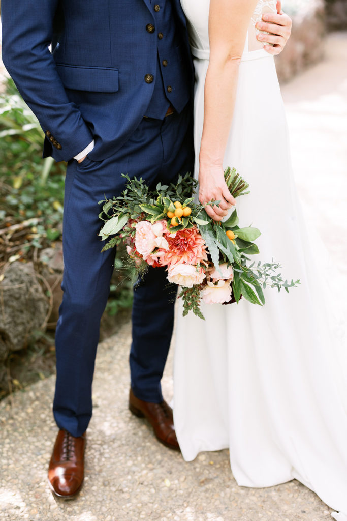 bride in spaghetti strap dress with lace back stands with groom in blue suit and red boutonniere 