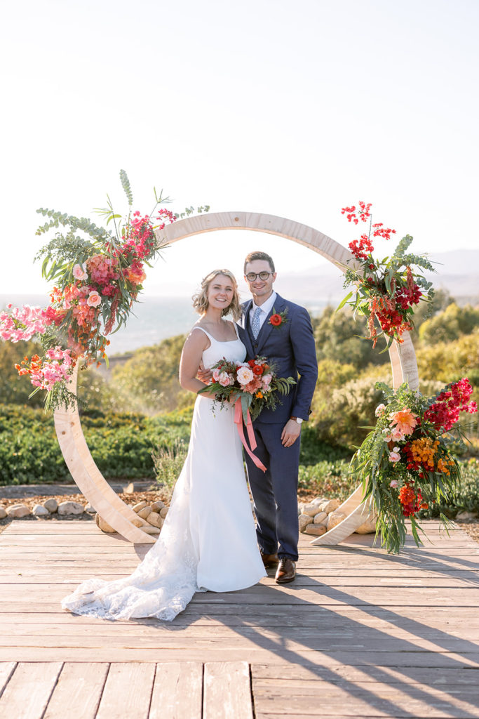 bride in spaghetti strap dress stands with groom in glasses and blue suit in front of their ceremony arch at Dos Pueblos Orchid Farm