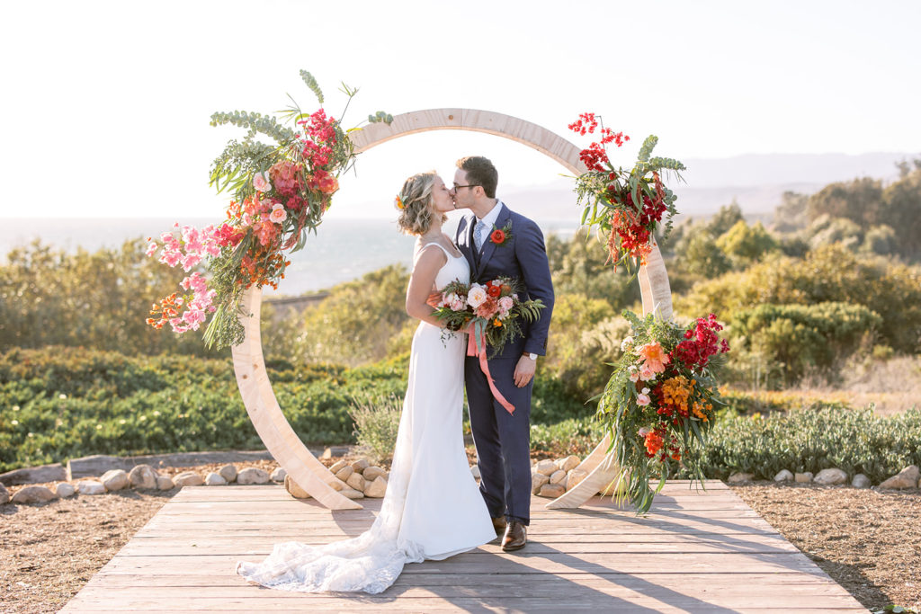 bride in spaghetti strap dress stands with groom in glasses and blue suit in front of their ceremony arch at Dos Pueblos Orchid Farm