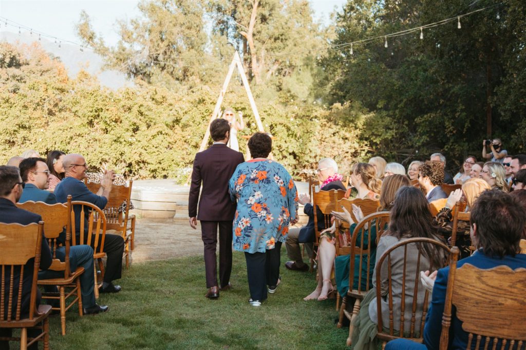 groom and mother of groom walk down aisle towards A-frame ceremony arch in retro inspired wedding in Ojai