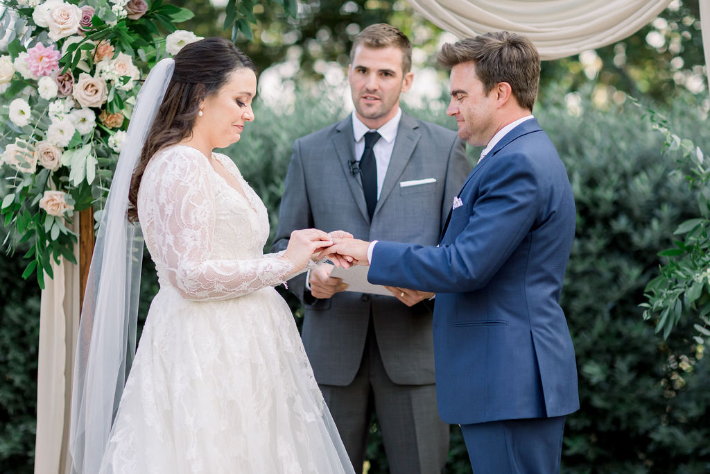 bride in long sleeve wedding dress exchanges rings with groom in blue suit during charming wedding ceremony at Maravilla Gardens