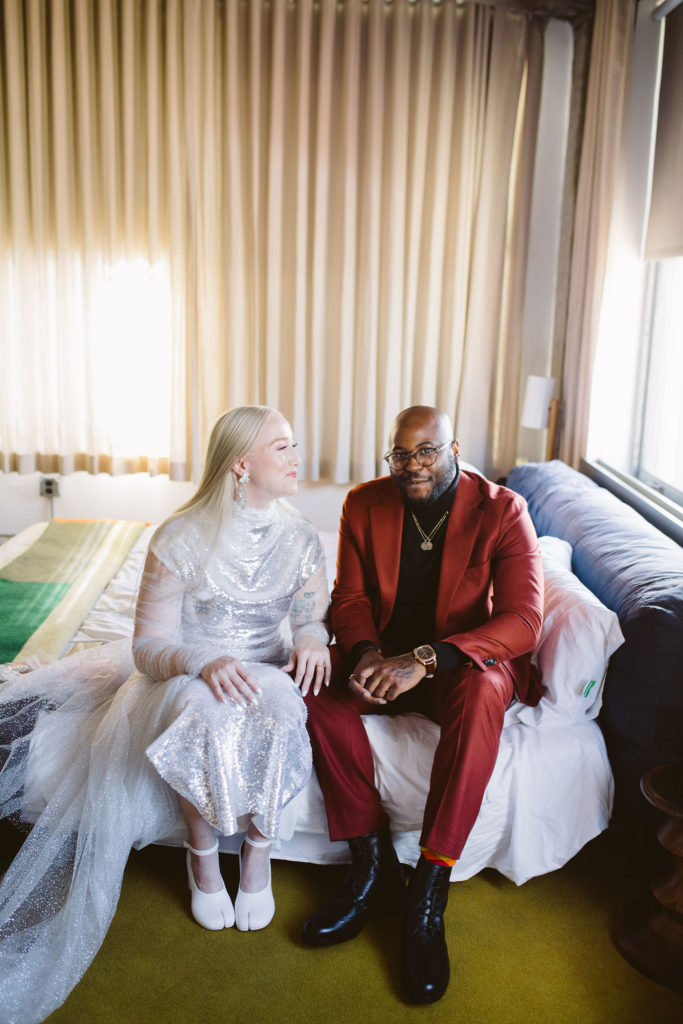 bride in silver wedding dress sits on bed with groom in red suit