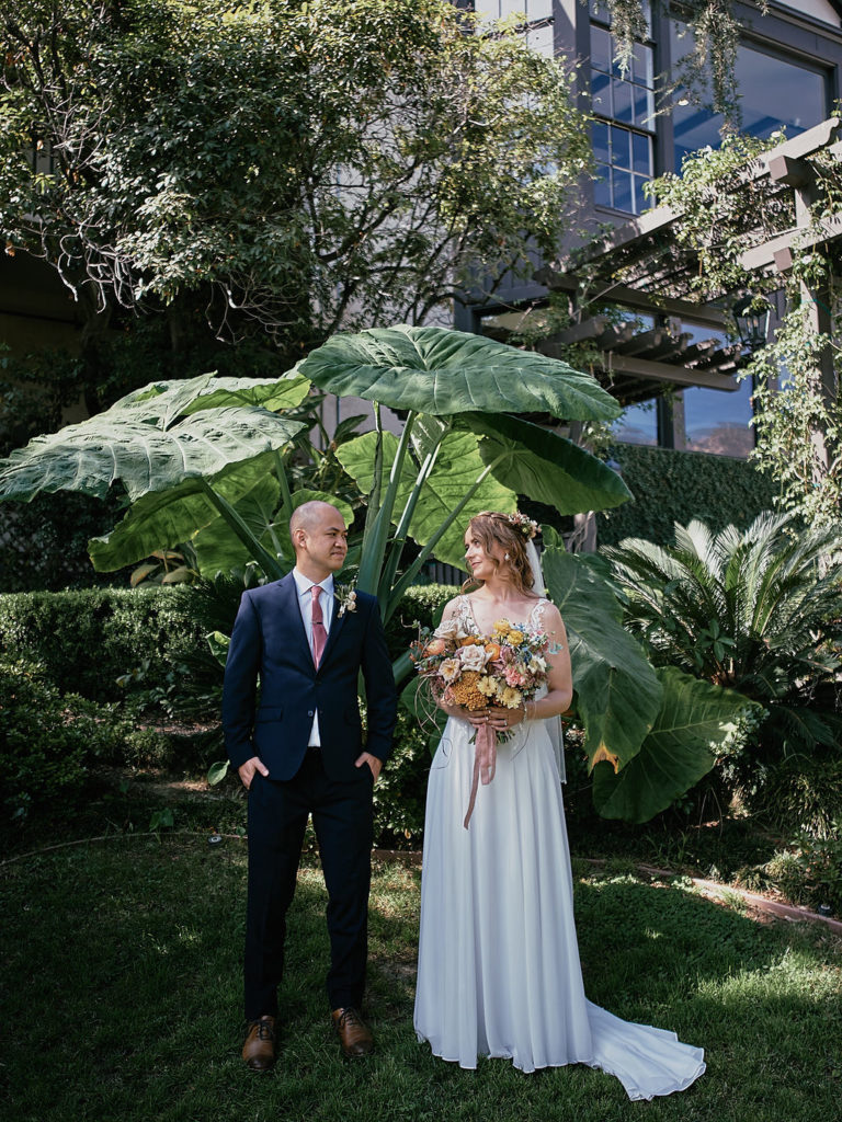 groom in navy suit and pink tie stands with bride in V-neck wedding dress and floral crown after their wedding a the Altadena Country Club