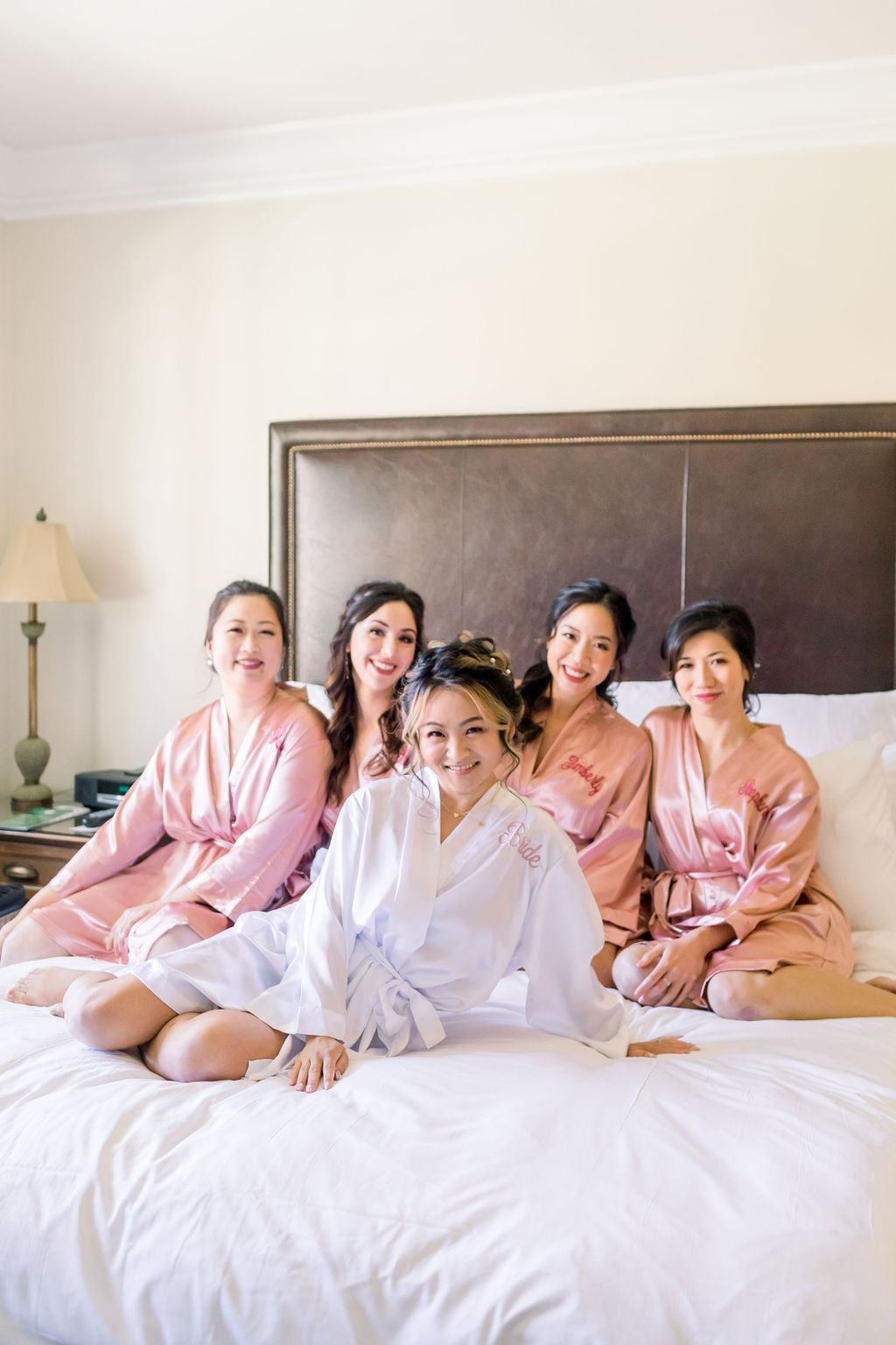 bride in white satin robe sits on bed with bridesmaids in personalized pink satin robes
