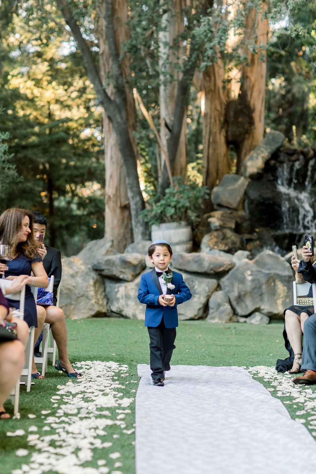 ring bearer in blue suit walks with ring box down aisle