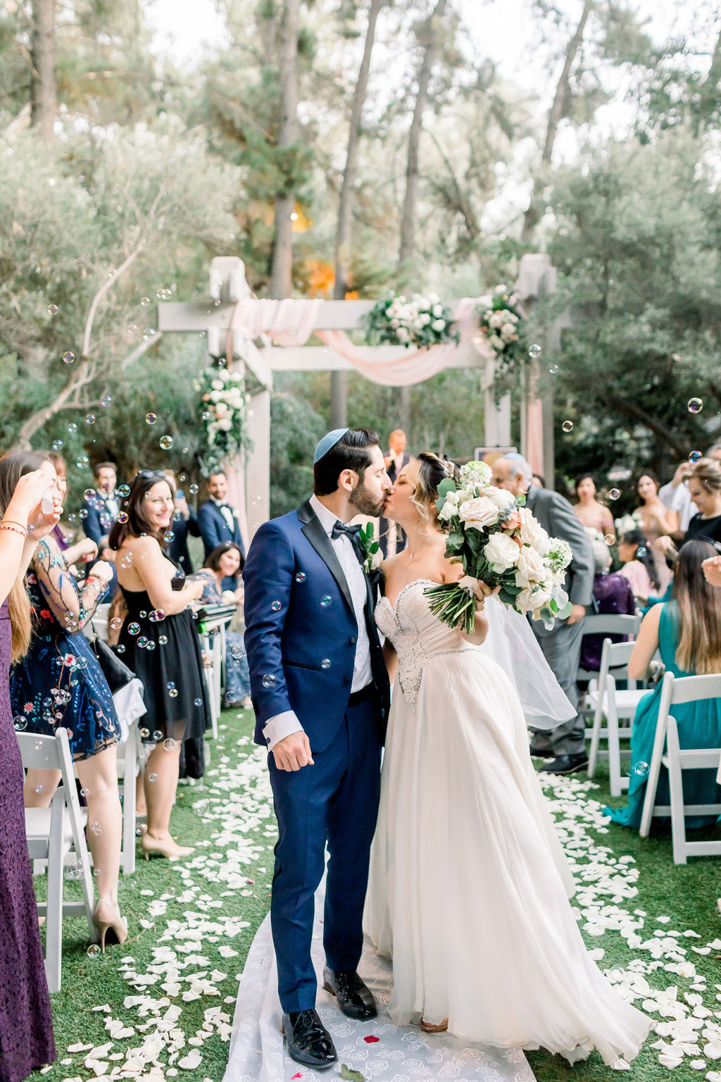 bride and groom kiss during recessional while guests blow bubbles at romantic and glamorous wedding ceremony at Calamigos Ranch