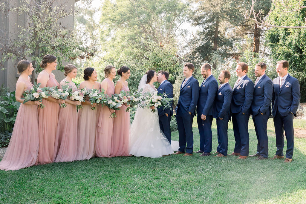 bride and groom kiss during wedding party photo with pink bridesmaid dresses and blue groomsmen suits with floral ties