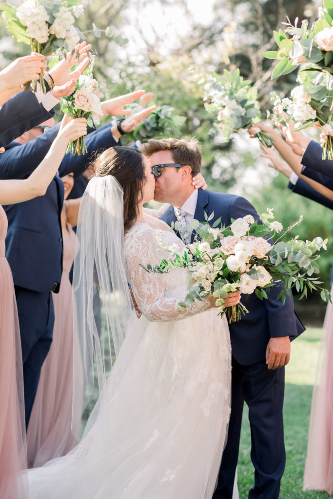bride in long sleeve wedding dress holds hands with groom in blue suit and floral tie while walking under a bouquet bridge