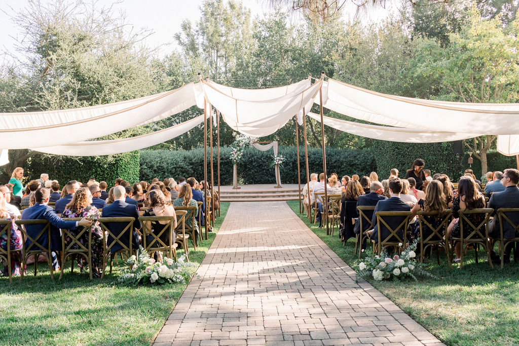 guests seated for charming wedding ceremony at Maravilla Gardens with soft pink chiffon drape and variety of pink florals with eucalyptus