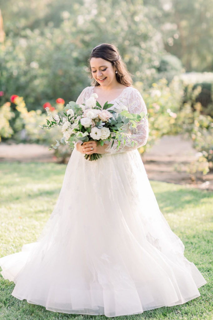 bride in long sleeve wedding dress with floral lace appliqué and sweetheart neckline stands with bridal bouquet of roses and eucalyptus