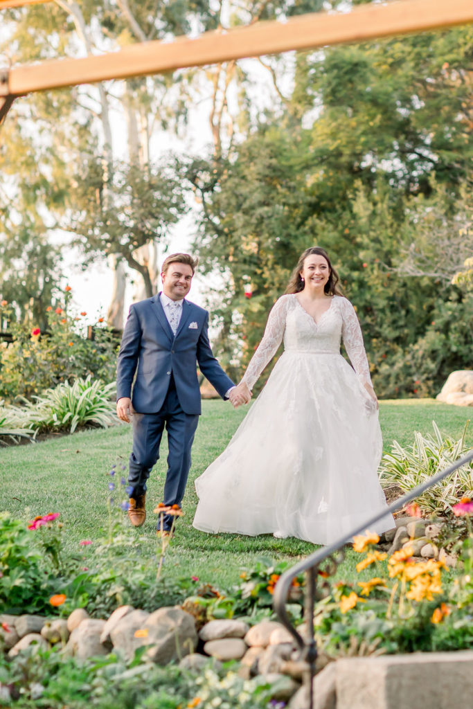 bride in long sleeve wedding dress walks with groom in blue suit towards reception at Maravilla Gardens