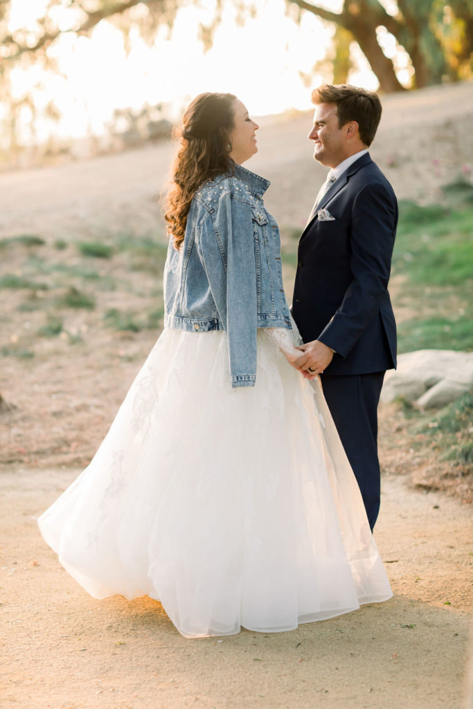 bride in long sleeve wedding dress with pearl embellished denim jacket walks with groom in blue suit and floral tie