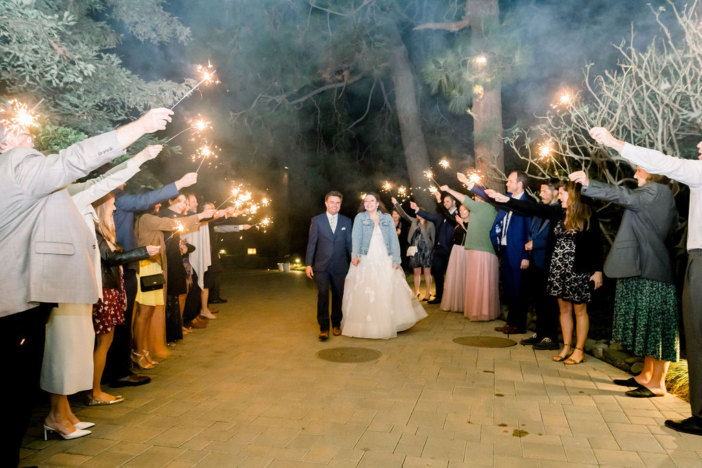 bride in long sleeve wedding dress with pearl embellished denim jacket walks with groom in blue suit and floral tie for grand exit with guests holding sparklers