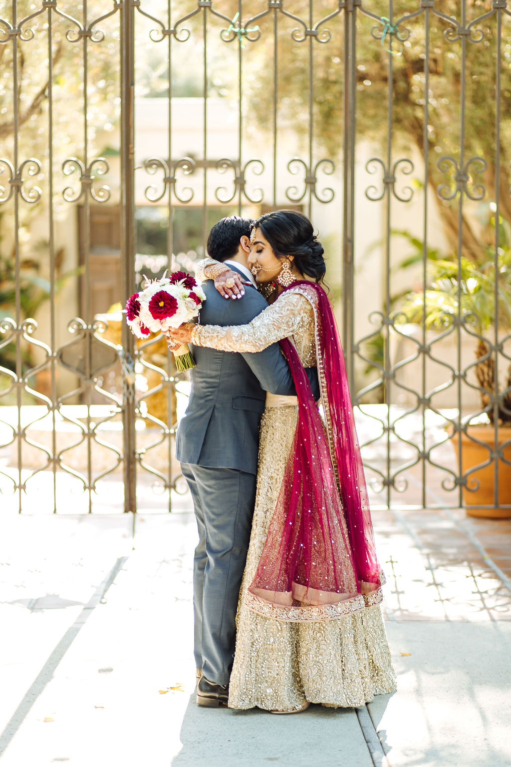 bride wearing embellished golden wedding saree and groom in dark grey suit with maroon tie embrace at Los Angeles River and Garden Center