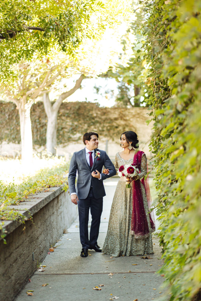 bride in golden wedding saree walks with groom in grey suit and maroon tie