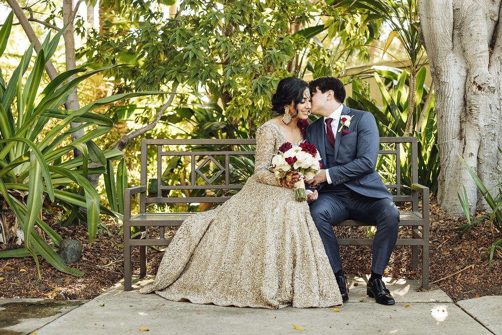 bride wearing embellished golden wedding saree and groom in dark grey suit with maroon sit on bench at Los Angeles River and Garden Center