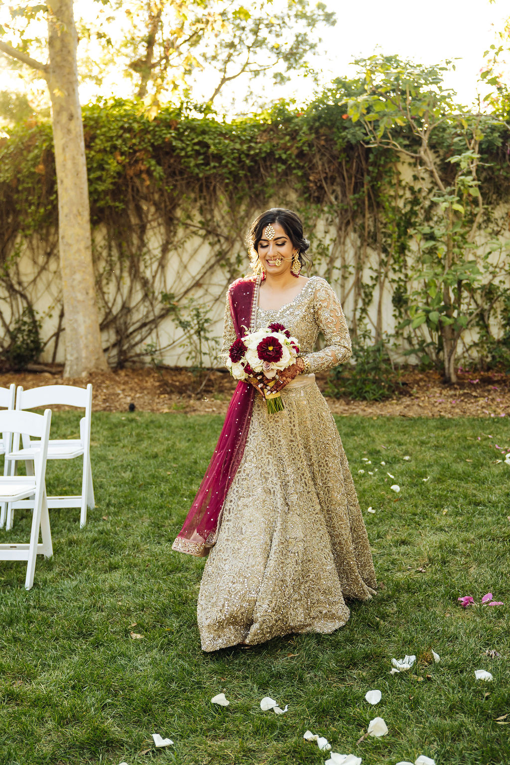 bride in embellished gold wedding saree and maroon sash walks down the aisle