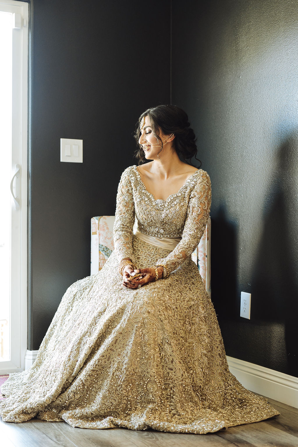 bride wearing embellished golden wedding saree, jewelry and henna sits in chair 