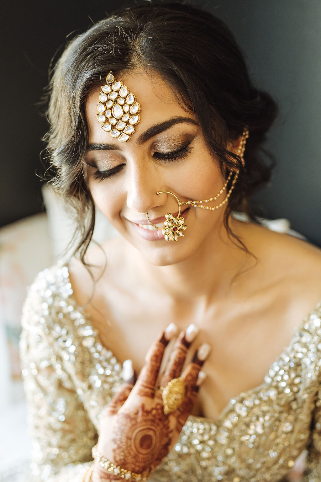 bride wearing embellished golden wedding saree, jewelry and henna sits in chair 