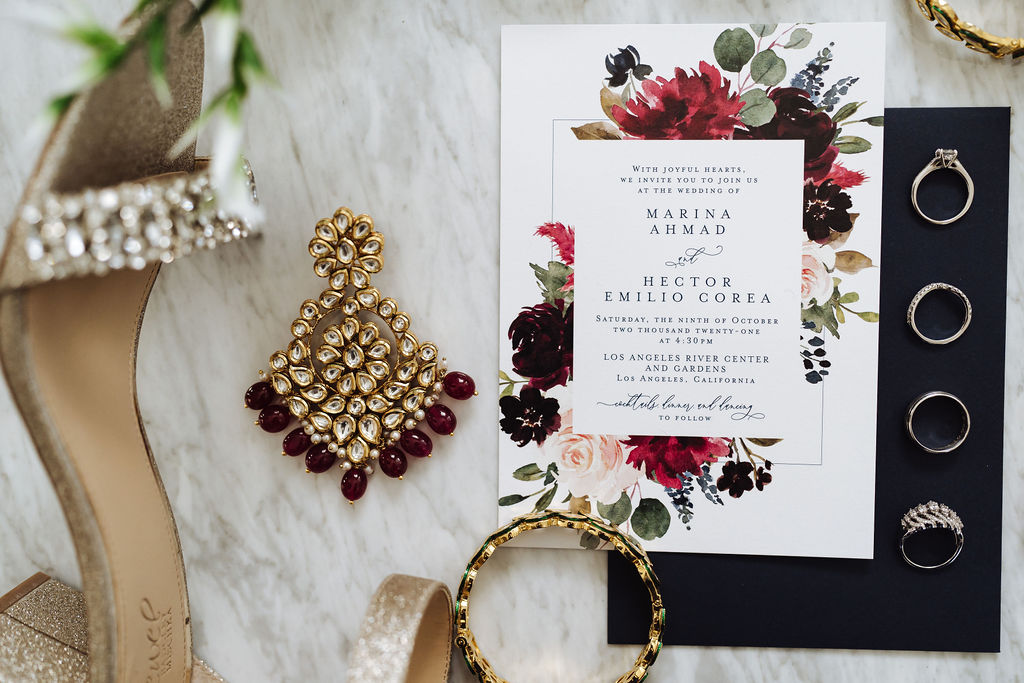 golden wedding invitation suite with maroon floral invitation and black envelope next to gold and maroon bridal jewelry
