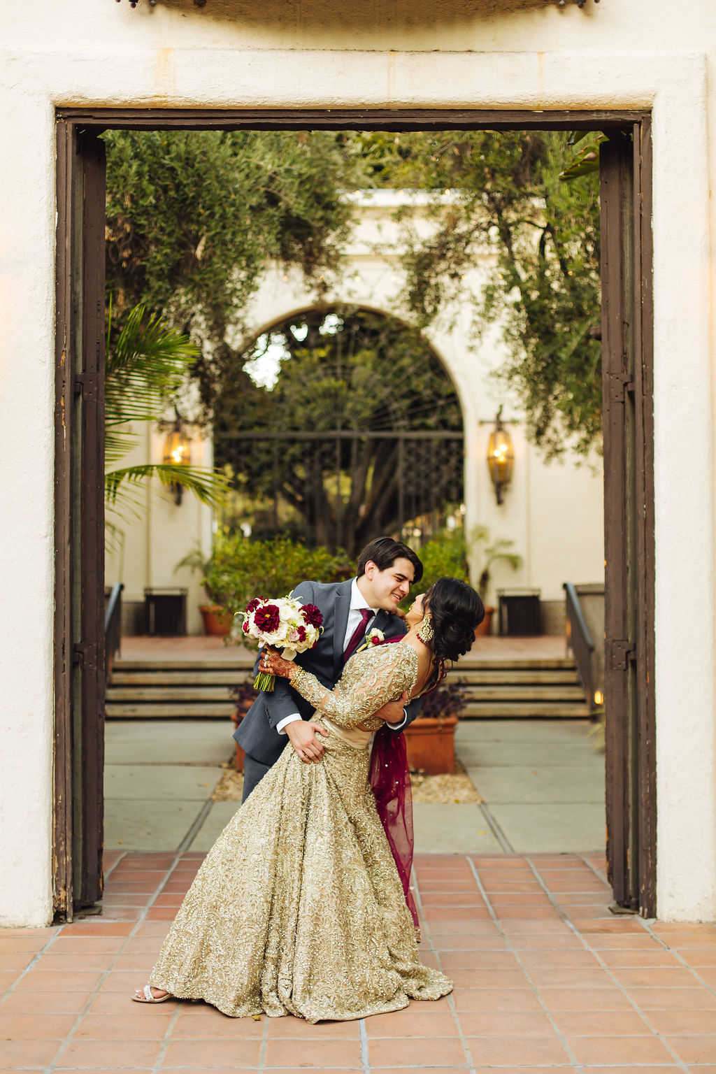 bride in embellished gold wedding saree with groom in dark grey suit and maroon tie take photos in the Los Angeles River Center and Gardens