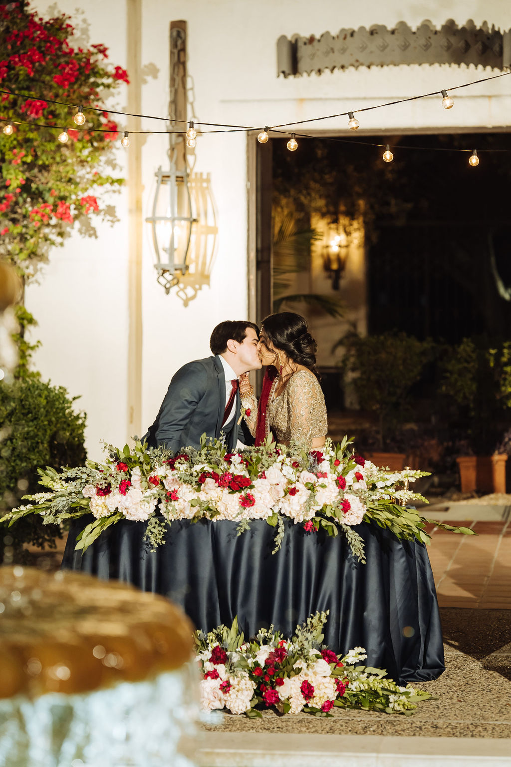 bride in embellished gold wedding saree with groom in dark grey suit and maroon tie kiss at the sweetheart table with silky black table cloth and large maroon floral arrangement