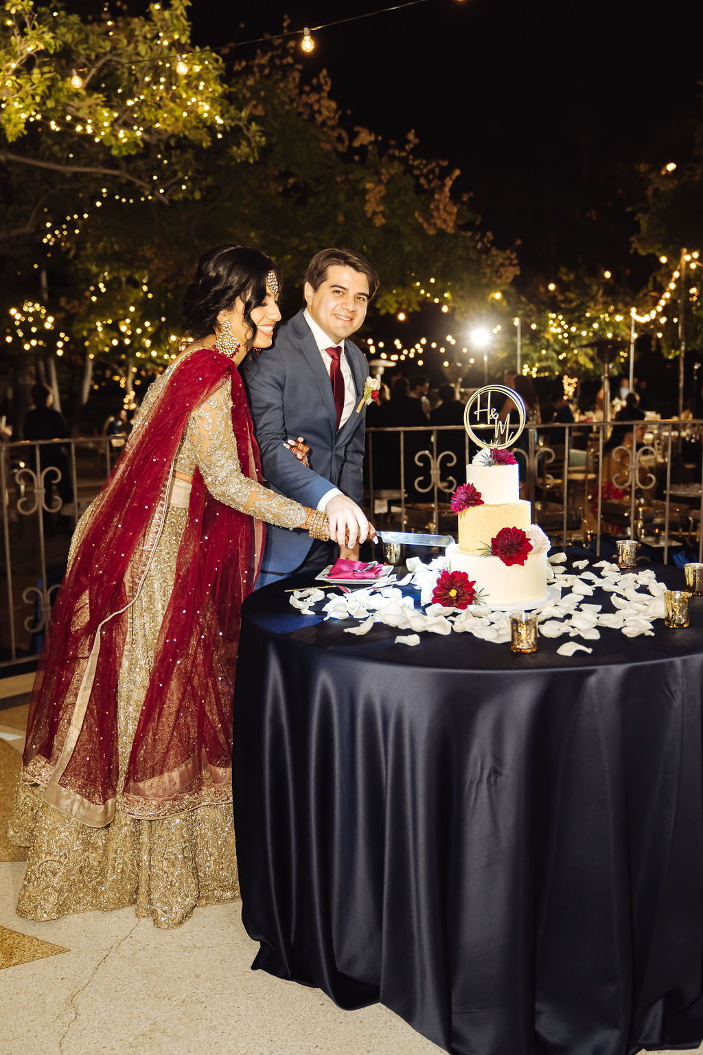 bride in embellished gold wedding saree with groom in dark grey suit and maroon tie cut wedding cake