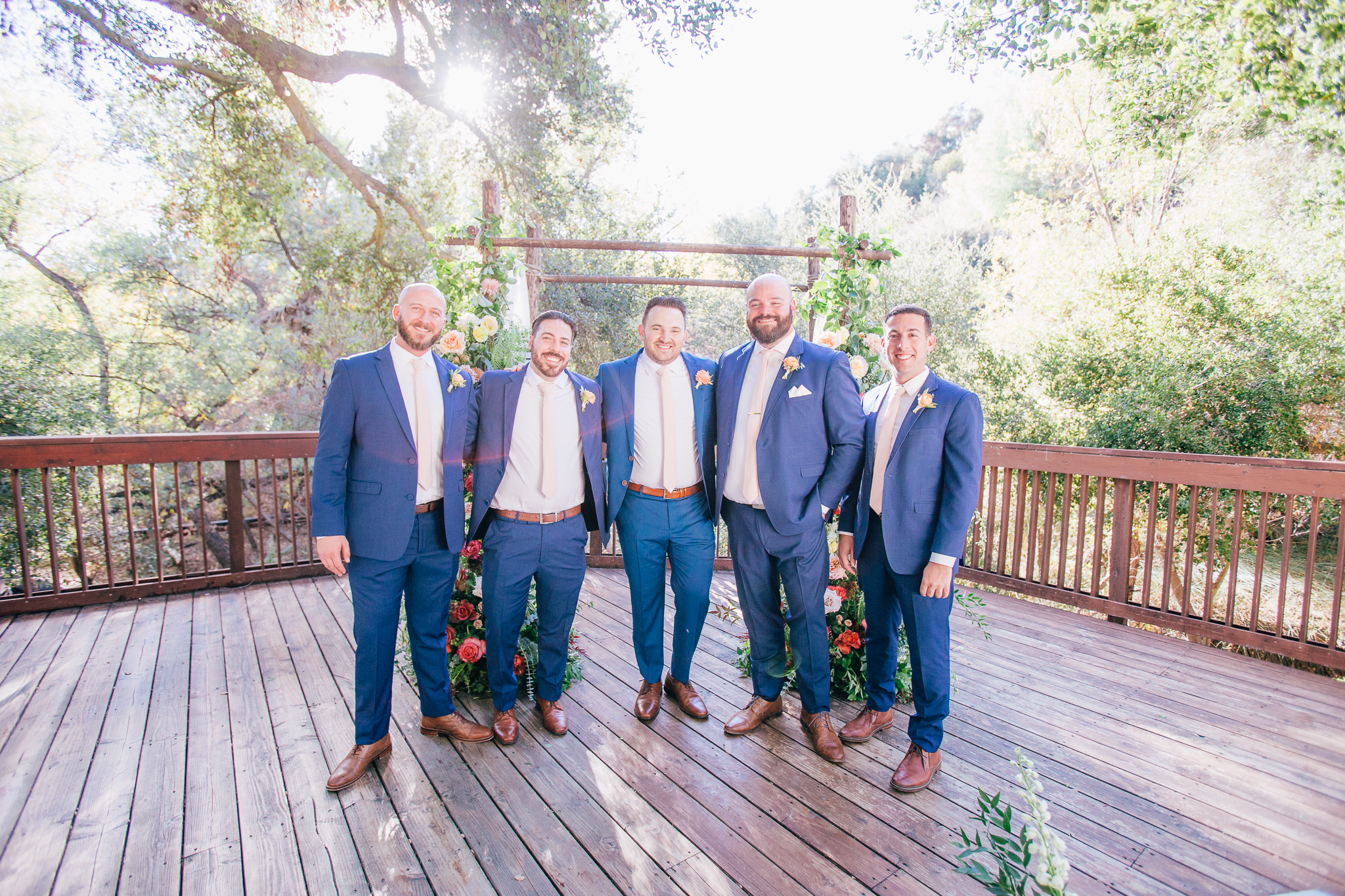 groom and groomsmen in blue suits with pink ties stand in front of wedding ceremony arch