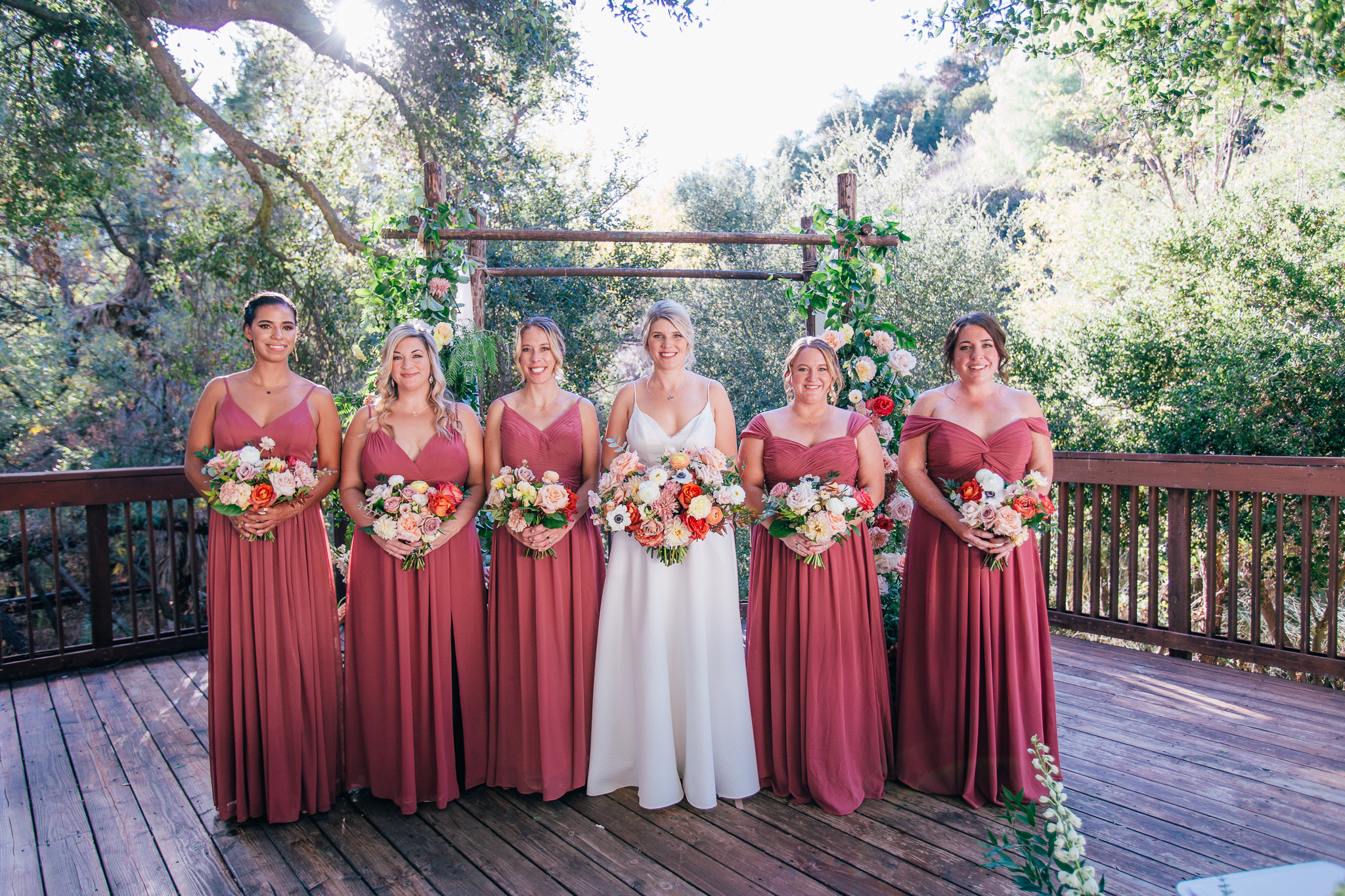 bride stands with bridesmaids in mauve dresses in front of wedding ceremony arch