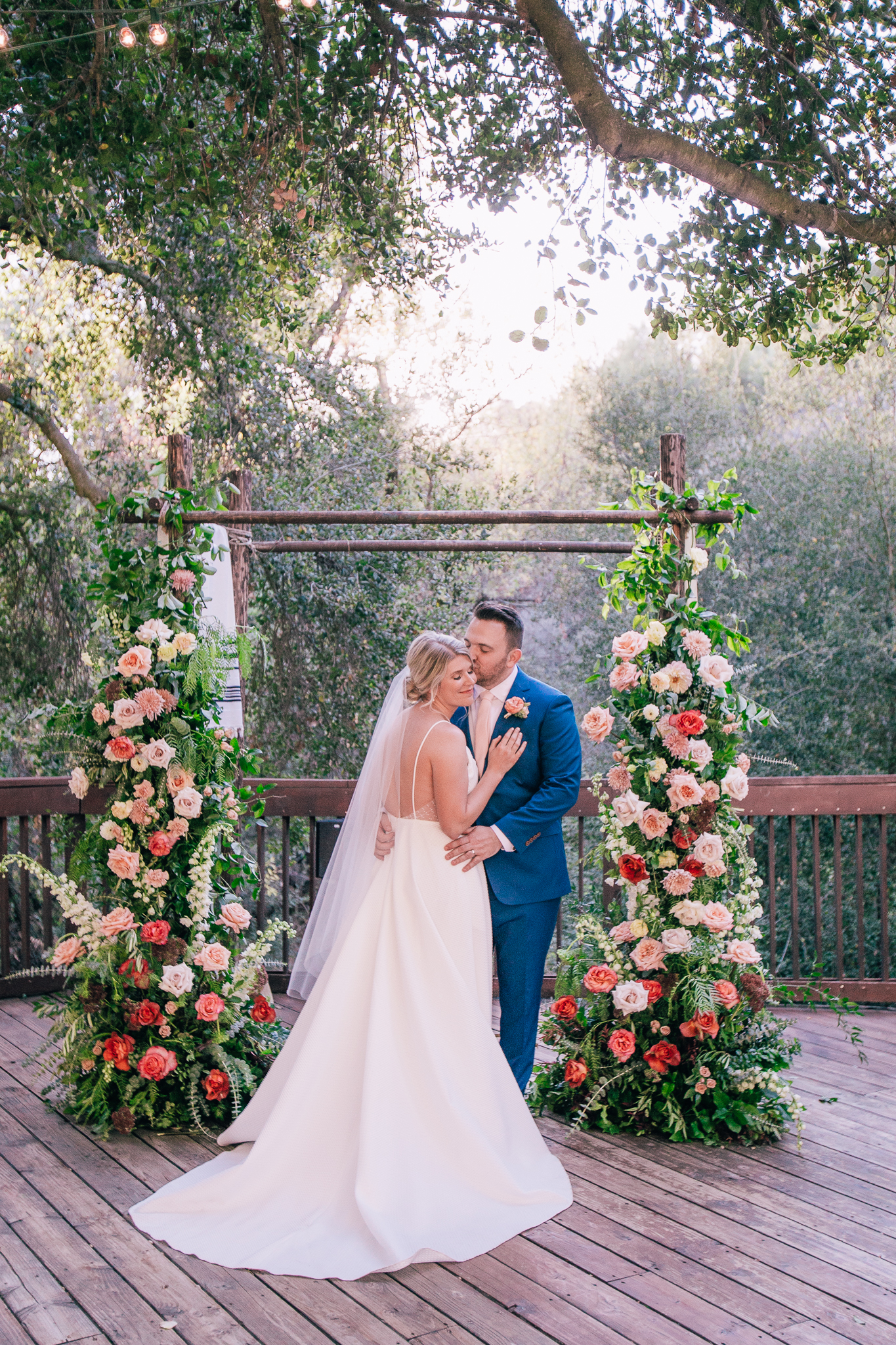 bride in spaghetti strap wedding dress stands with groom in blue suit and light pink tie in front of their wedding ceremony arch with bright florals
