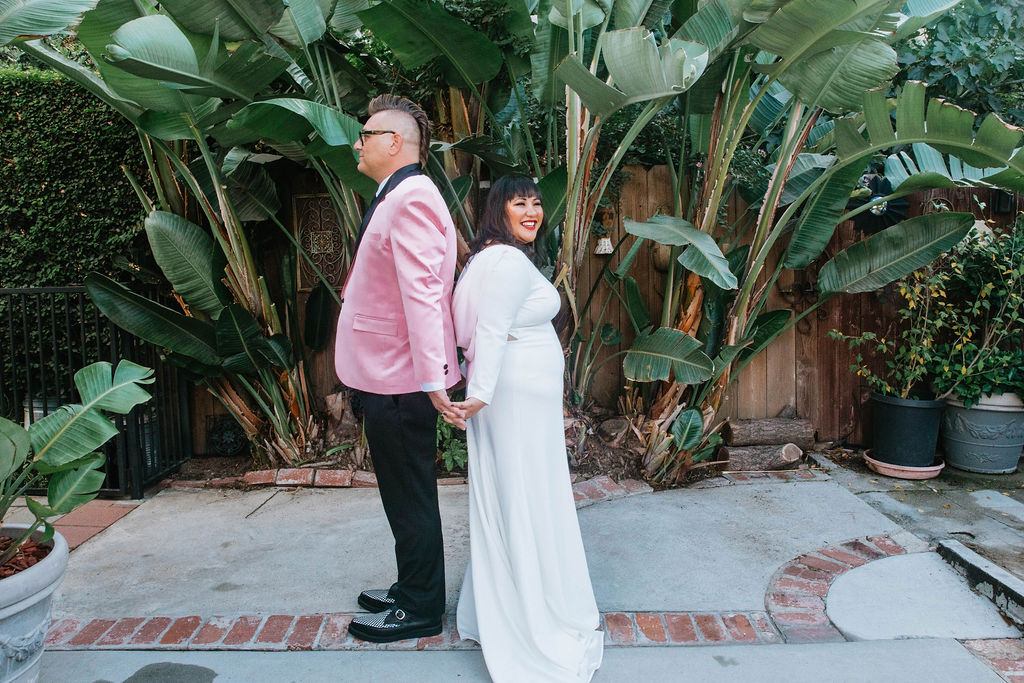 bride in long sleeve deep v-neck wedding dress stands back to back with groom in pink suit jacket and houndstooth shoes during first look 