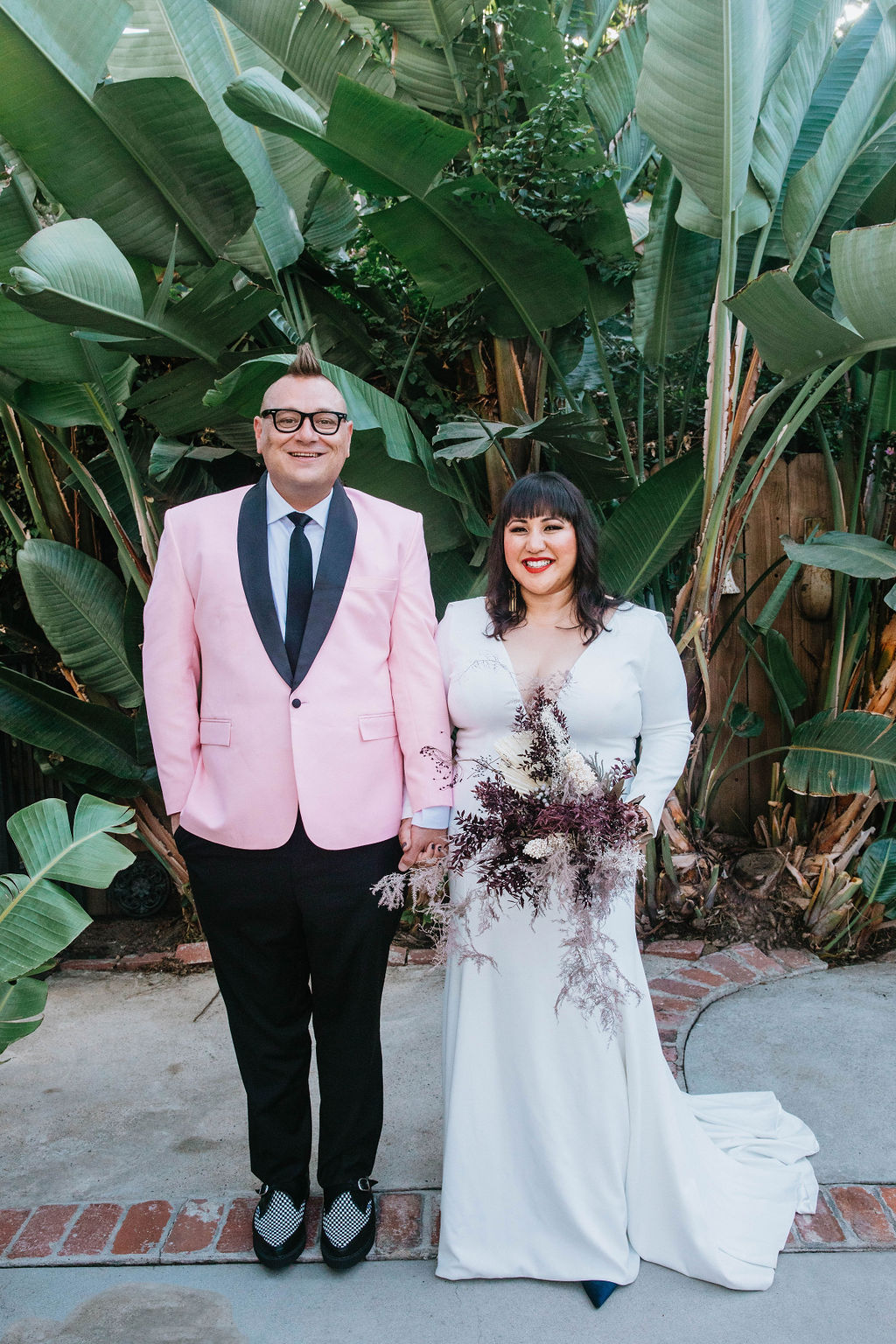 bride in long sleeve deep v-neck wedding dress holds bouquet of dried florals with groom in pink suit jacket and houndstooth shoes 