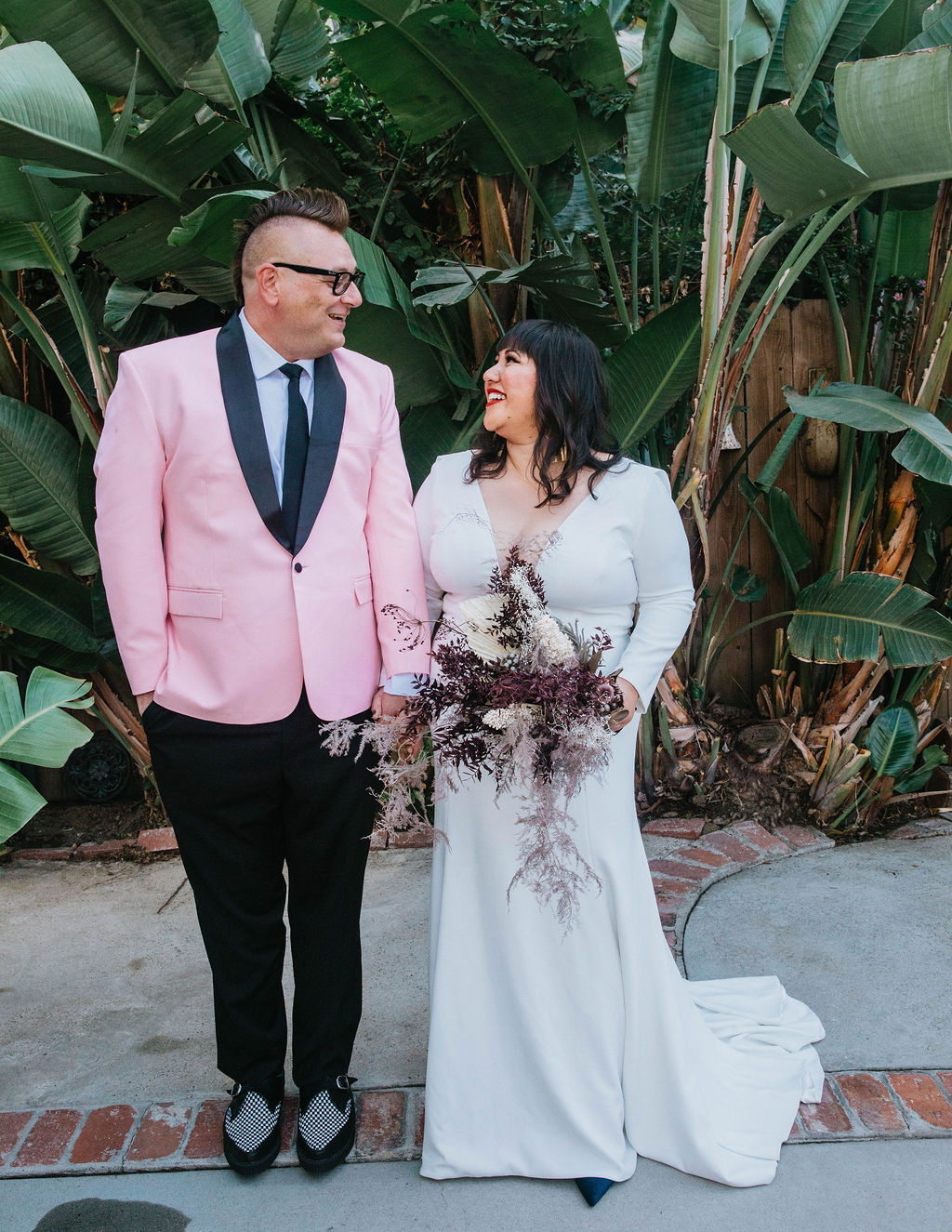 bride in long sleeve deep v-neck wedding dress holds bouquet of dried florals with groom in pink suit jacket and houndstooth shoes 