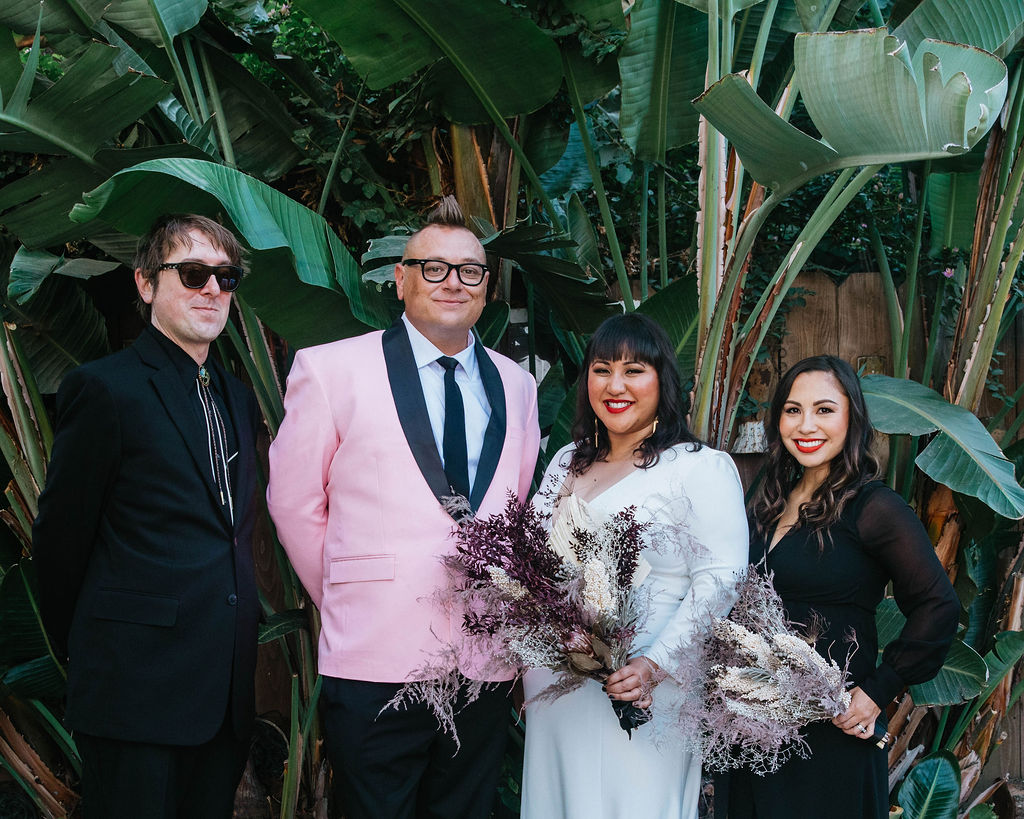 bride in long sleeve deep v-neck wedding dress holds bouquet of dried florals with groom in pink suit jacket and houndstooth shoes stand with with their wedding party in black suit and dress