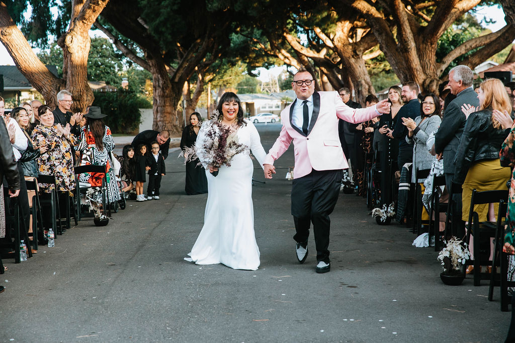bride in long sleeve deep v-neck wedding dress celebrates with groom in pink suit jacket after wedding ceremony under an urban canopy of trees in the middle of a residential street in Long Beach with dried floral aisle arrangements and black chairs