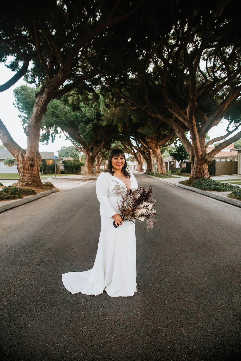 bride in long sleeve deep v-neck wedding dress holding bouquet of purple dried florals stands at ceremony site in the middle of a residential street under an urban canopy of trees