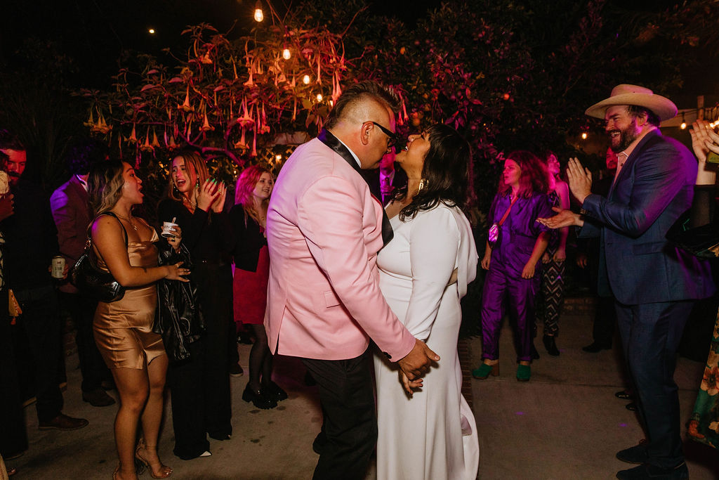 bride in long sleeve deep v-neck wedding dress and groom in pink suit jacket have first dance in backyard wedding reception