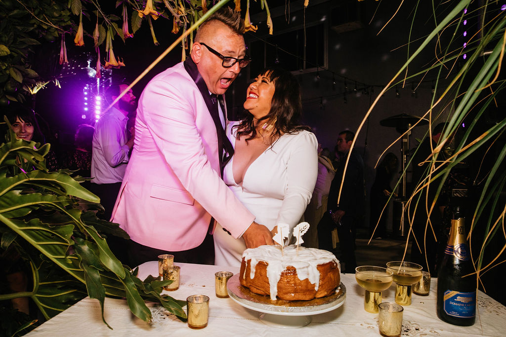 bride in long sleeve deep v-neck wedding dress cuts cake with groom in pink suit jacket 