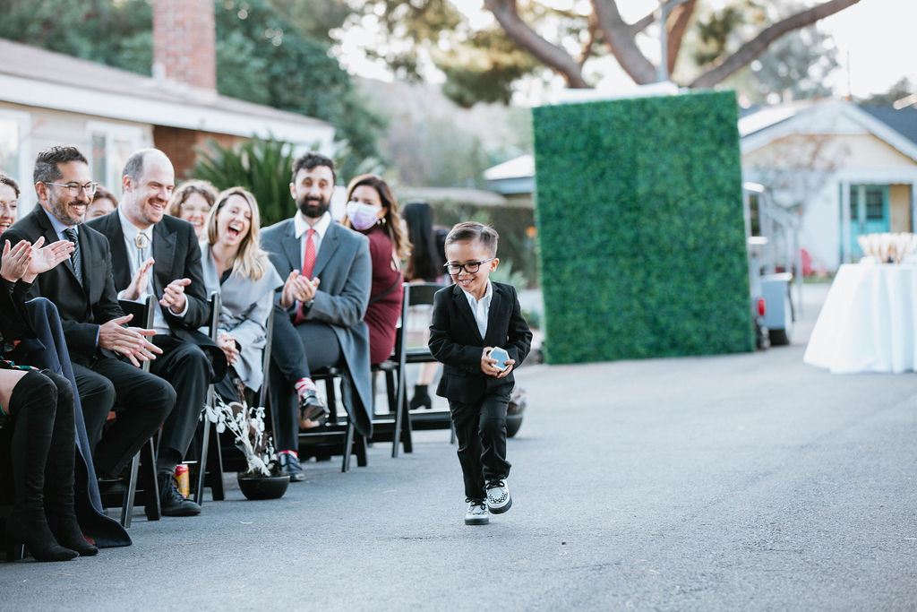 ring bearer in black suit runs down the wedding aisle with ring box in hand