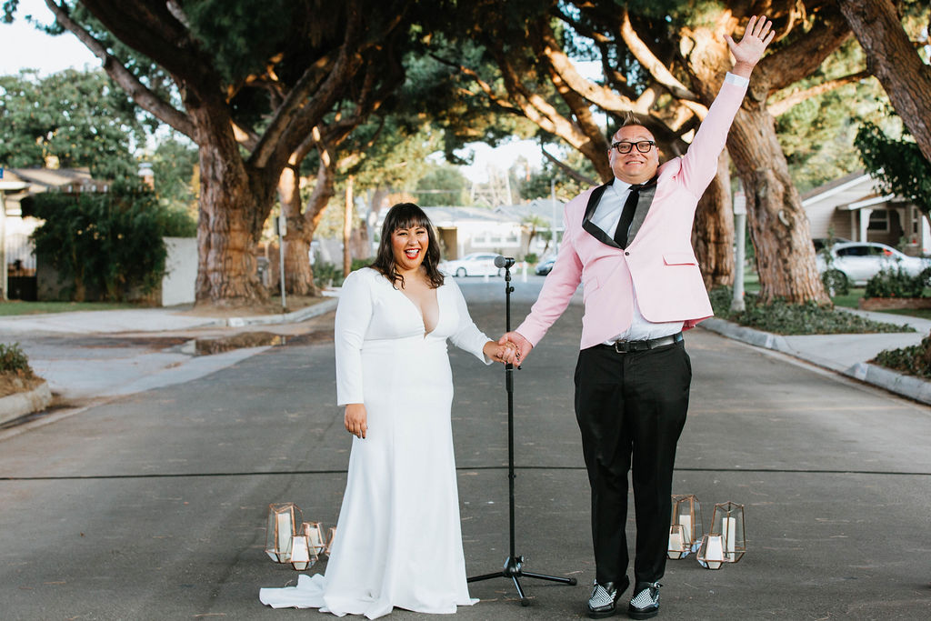 bride in long sleeve deep v-neck wedding dress celebrates with groom in pink suit jacket after wedding ceremony under an urban canopy of trees in the middle of a residential street in Long Beach with dried floral aisle arrangements and black chairs