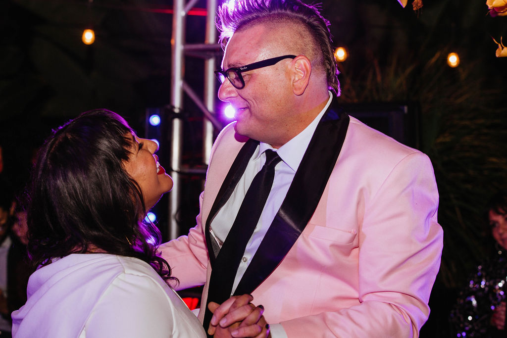 bride in long sleeve deep v-neck wedding dress and groom in pink suit jacket have first dance in backyard wedding reception