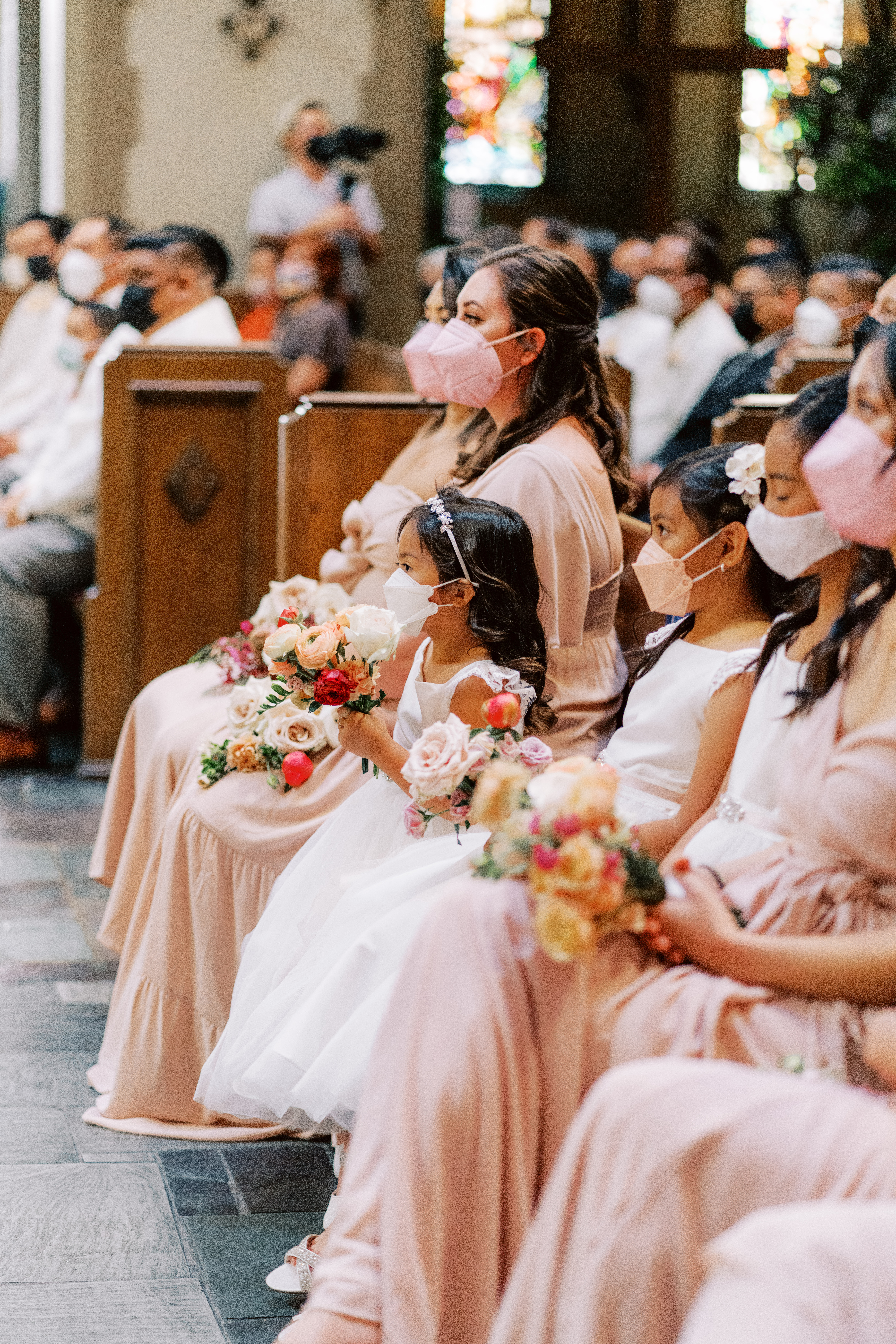 bridesmaids in light peach dresses with m matching masks sit with flower girls in white dresses and masks during church wedding ceremony