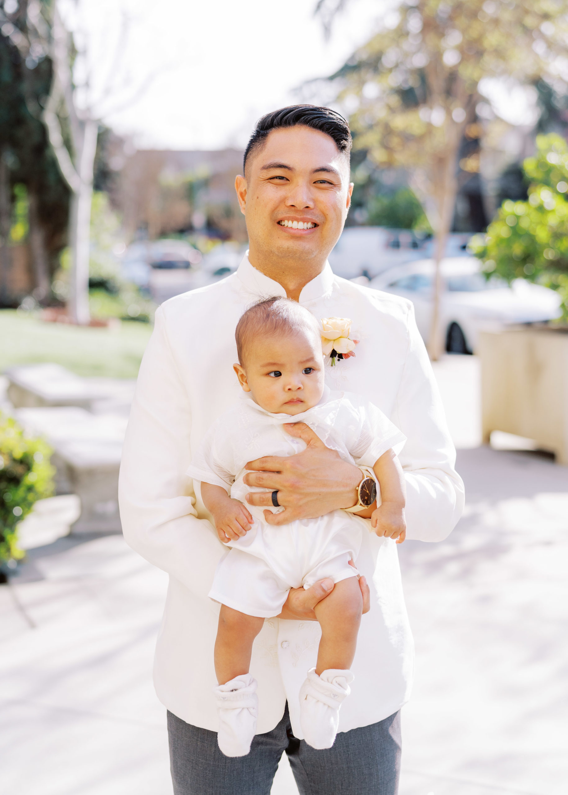 groom in traditional white wedding attire holds baby during wedding portraits 
