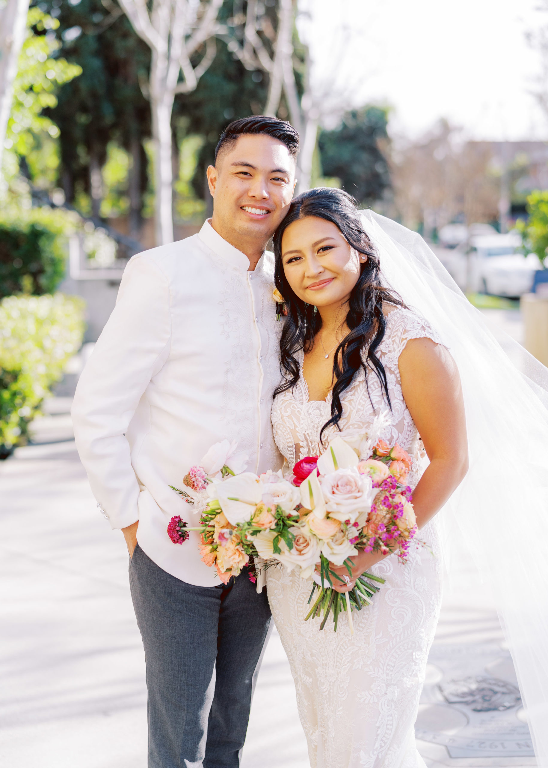 bride in lace wedding dress and tropical floral bouquet stands with groom in white wedding attire outside of church 