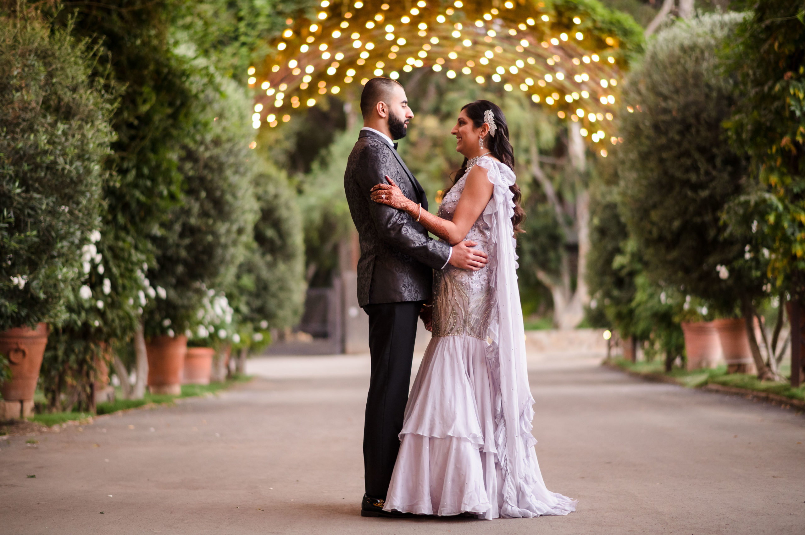 bride in lilac trumpet gown with crystals and groom in charcoal black textured suit jacket stand under canopy of lights