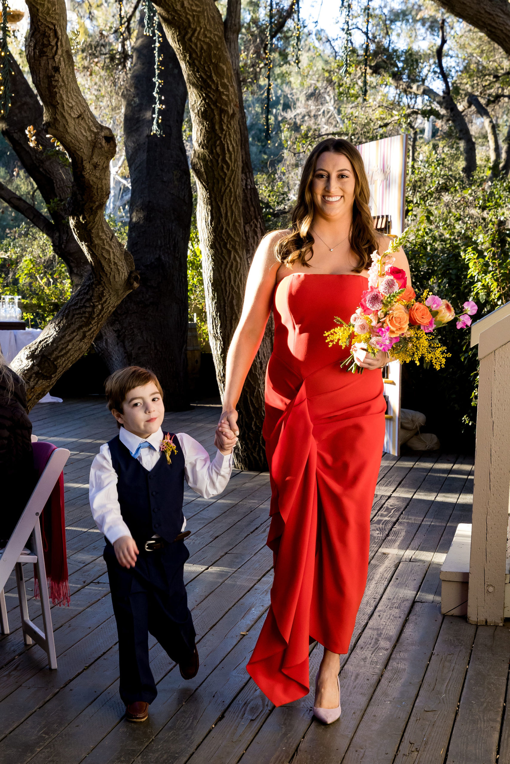 bridesmaid in red dress and bright floral bouquet holds hand of young ring bearer wearing blue vest and bow tie