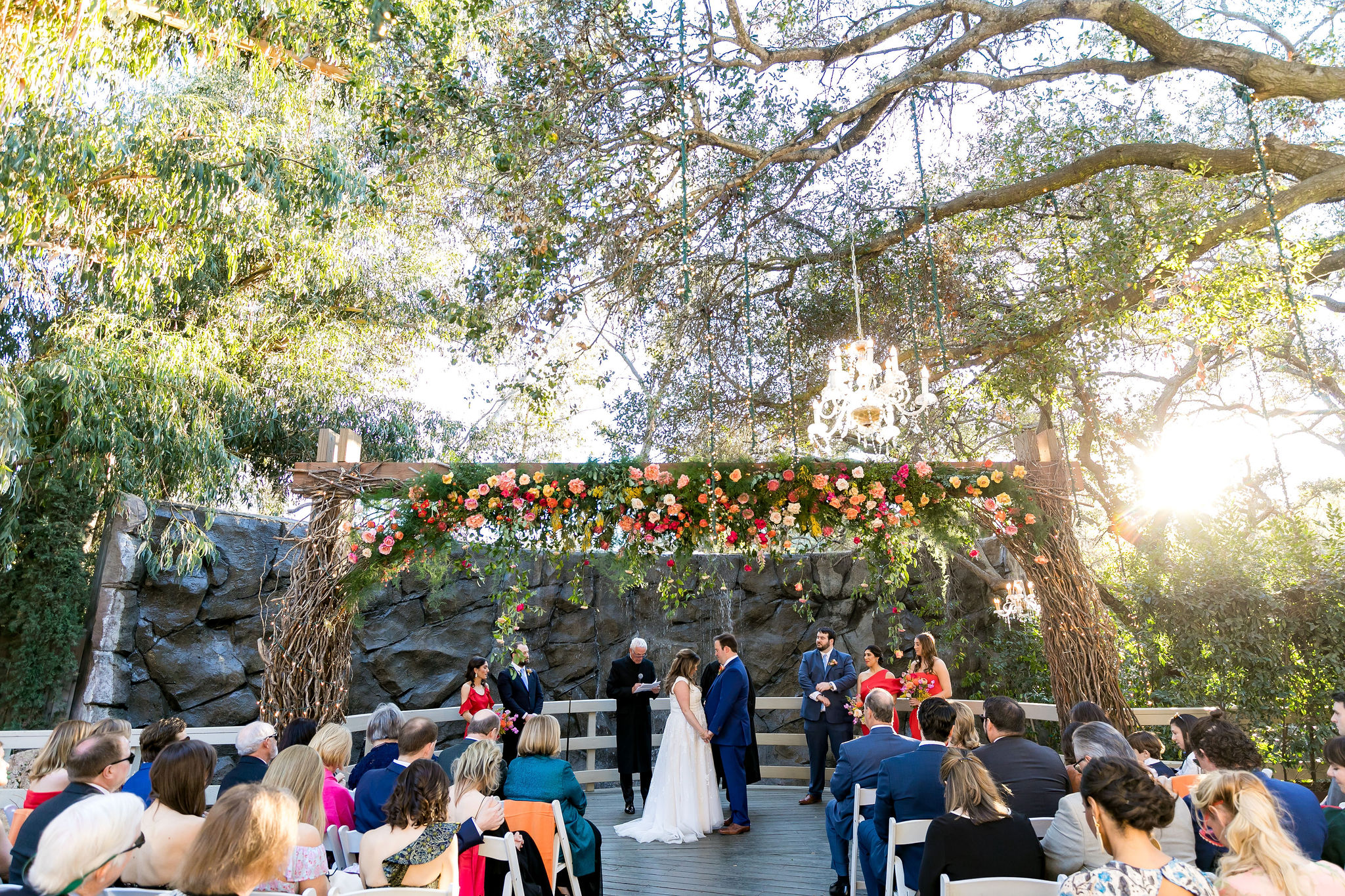 bright and energetic wedding ceremony at the Oak Room at Calamigos Ranch with bright red, pink, orange and white florals hanging in front of waterfall