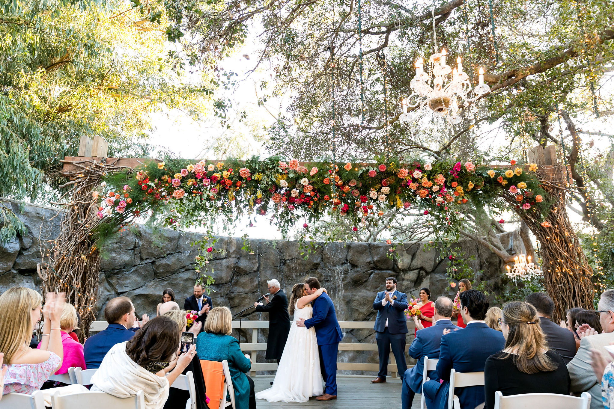 bride and groom first kiss during bright and energetic wedding ceremony at Calamigos Ranch with red, pink, orange, yellow, purple and white florals hanging in front of waterfall 